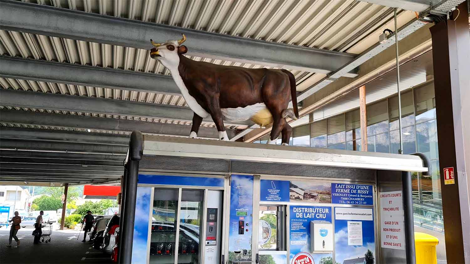 A distributeur de lait cru installed in a supermarket car park canopy in La Ravoire, Auvergne-Rhône-Alpes, France, with a life-size fibreglass cow on top and 24-hour operation signage