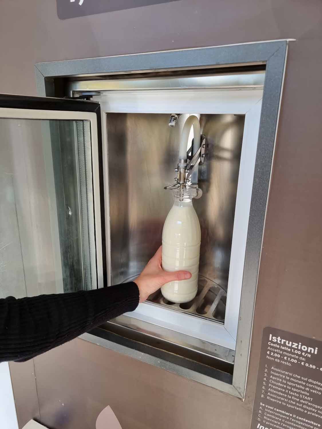 A hand holding a glass bottle being filled at an Italian distributore di latte crudo, with the instruction panel showing a price of €1.00 per liter