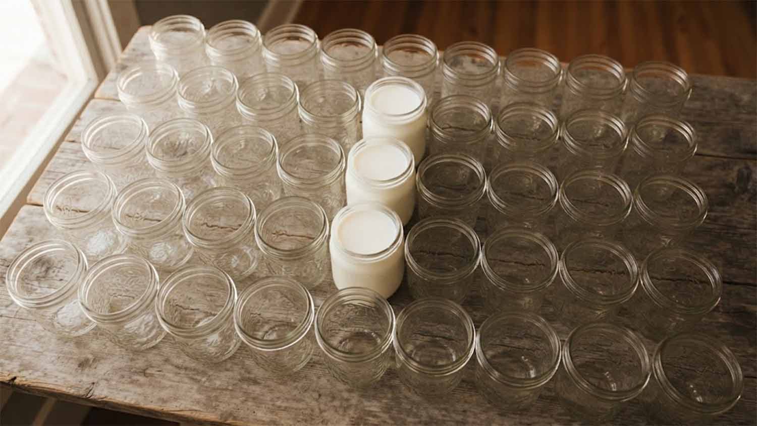 Three glass jars filled with fresh raw milk among dozens of empty mason jars on a weathered wooden table, lit by soft window light.