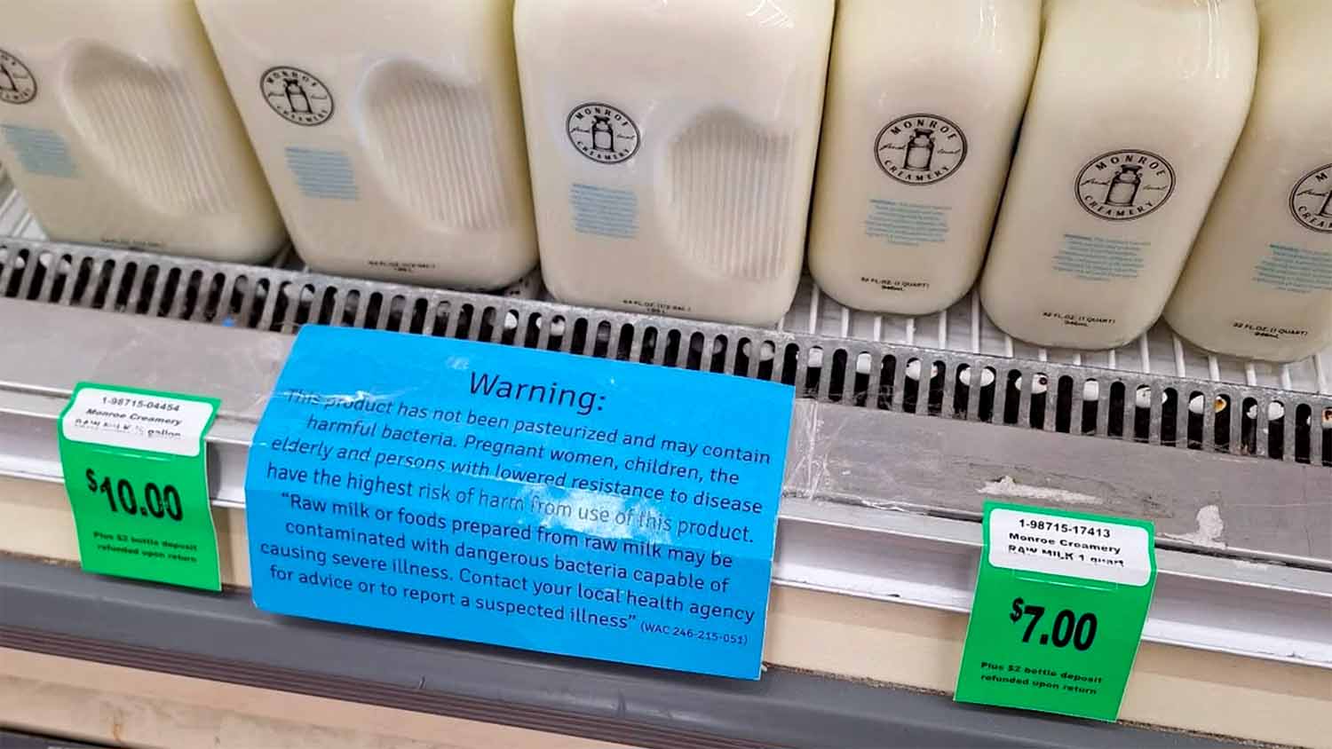 Glass bottles of raw milk on a store shelf, with a state-mandated warning label reading 'This product has not been pasteurized and may contain harmful bacteria.'